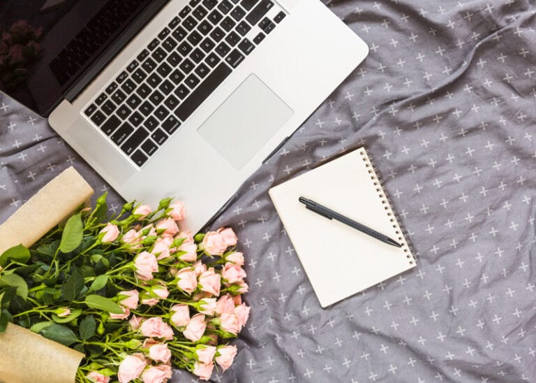 Ordinateur portable avec un carnet et un stylo sur un lit, entouré de roses – illustration d’un espace de travail créatif pour les créateurs de site web.