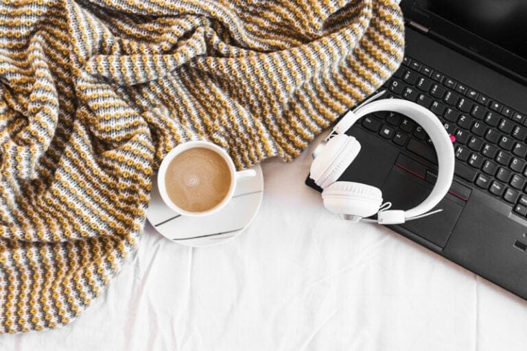 Ordinateur portable noir avec un casque audio blanc, une tasse de café et un plaid cosy, représentant l'organisation d'un freelance.