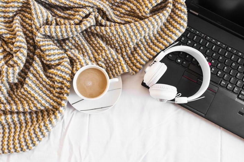 Ordinateur portable noir avec un casque audio blanc, une tasse de café et un plaid cosy, représentant l'organisation d'un freelance.