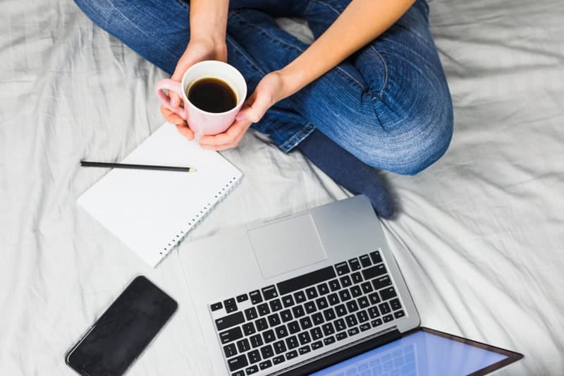 Personne assise en tailleur avec une tasse de café, carnet, ordinateur portable et téléphone, ambiance cosy propice à l’écriture