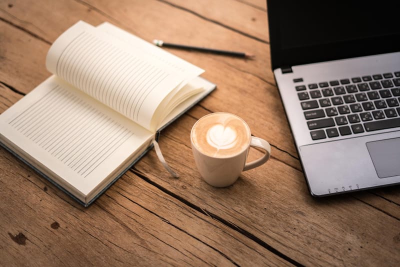 Tasse de café latte posée entre un carnet ouvert et un ordinateur portable sur une table en bois – ambiance écriture et blog littéraire.