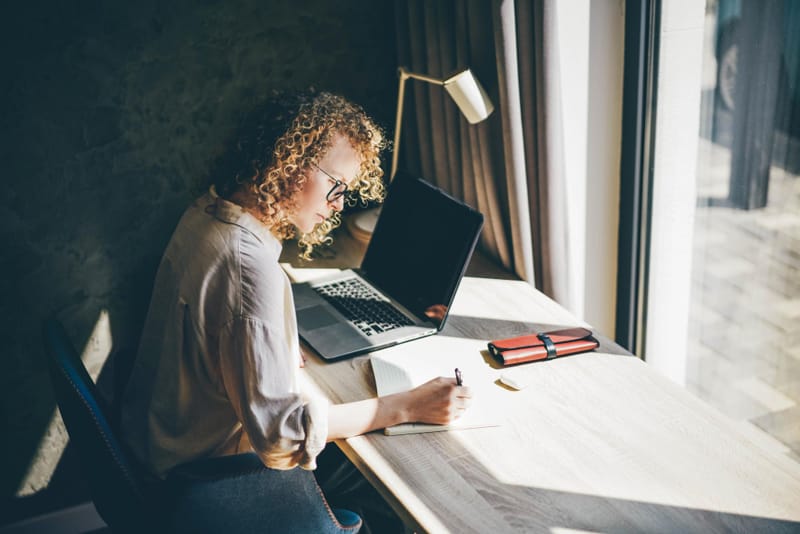 Personne concentrée en train d’écrire dans un carnet, devant un ordinateur portable, assise à un bureau lumineux.