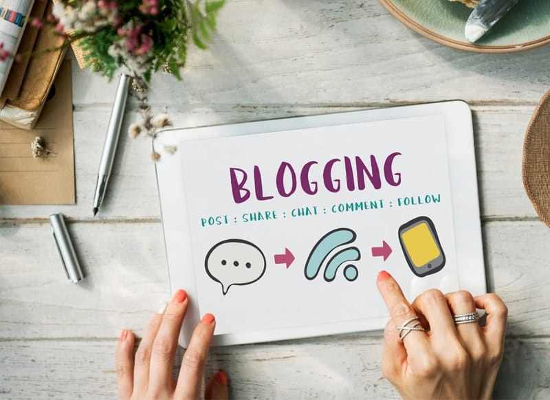 Tablette affichant le mot "Blogging" entouré d’icônes de partage et de communication, avec des mains prêtes à interagir.